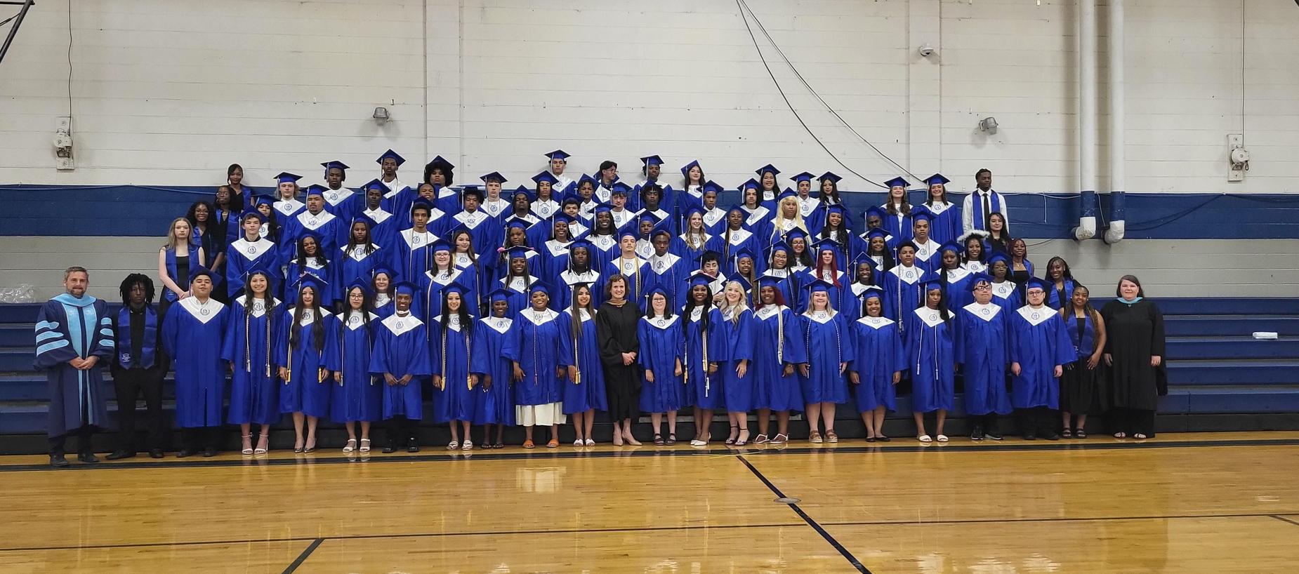 Large group photo of graduates in blue gowns with teachers on the bleachers.