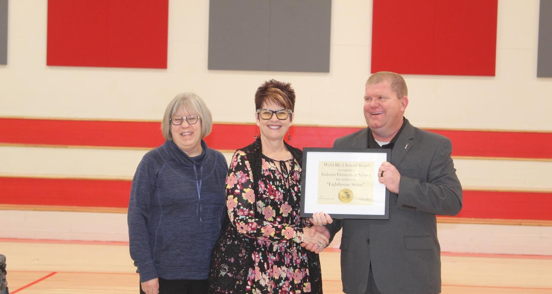 Three adults presenting an award certificate in a gym setting, smiling at the camera.