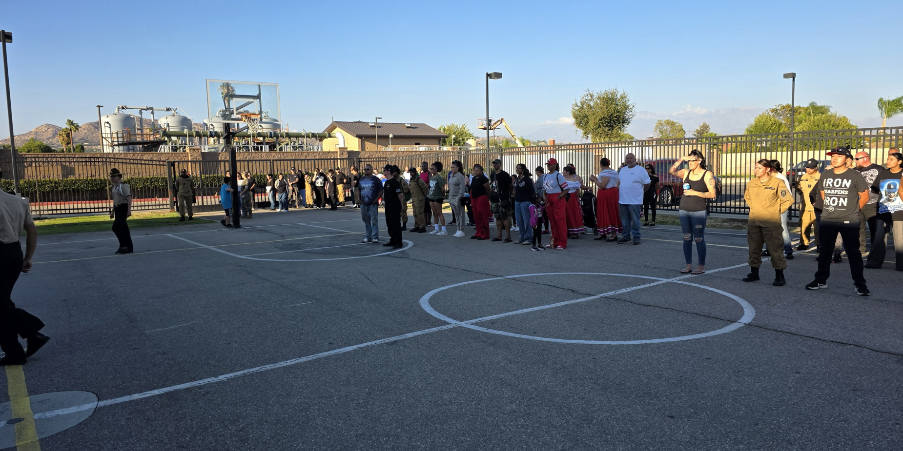 A group of people waiting in line on a basketball court near a fence and buildings.