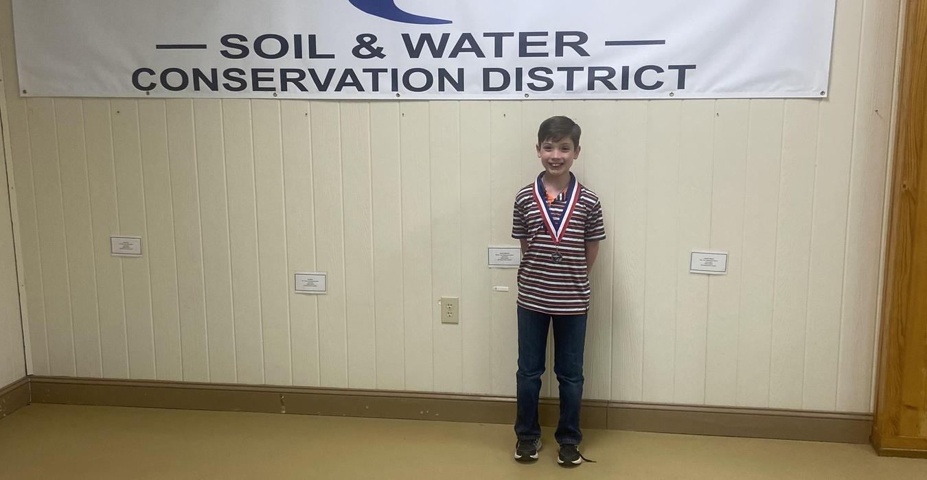 Child wearing a medal stands in front of a conservation district banner.