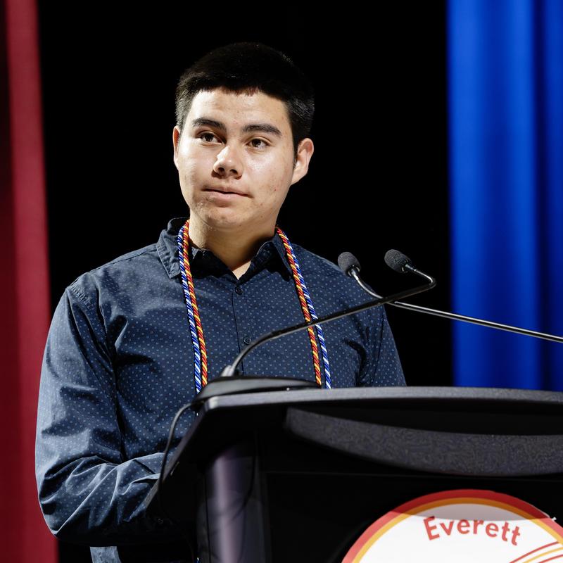 A young man wearing a dark shirt stands at a podium, speaking at an event.