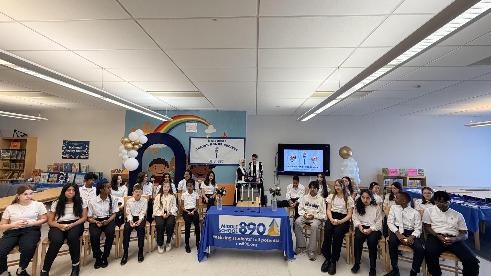 Students seated in a school library with a presentation setup and decorations.