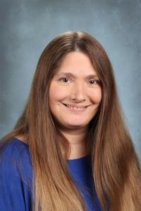 Woman with long brown hair in a blue shirt smiling against a gray background.