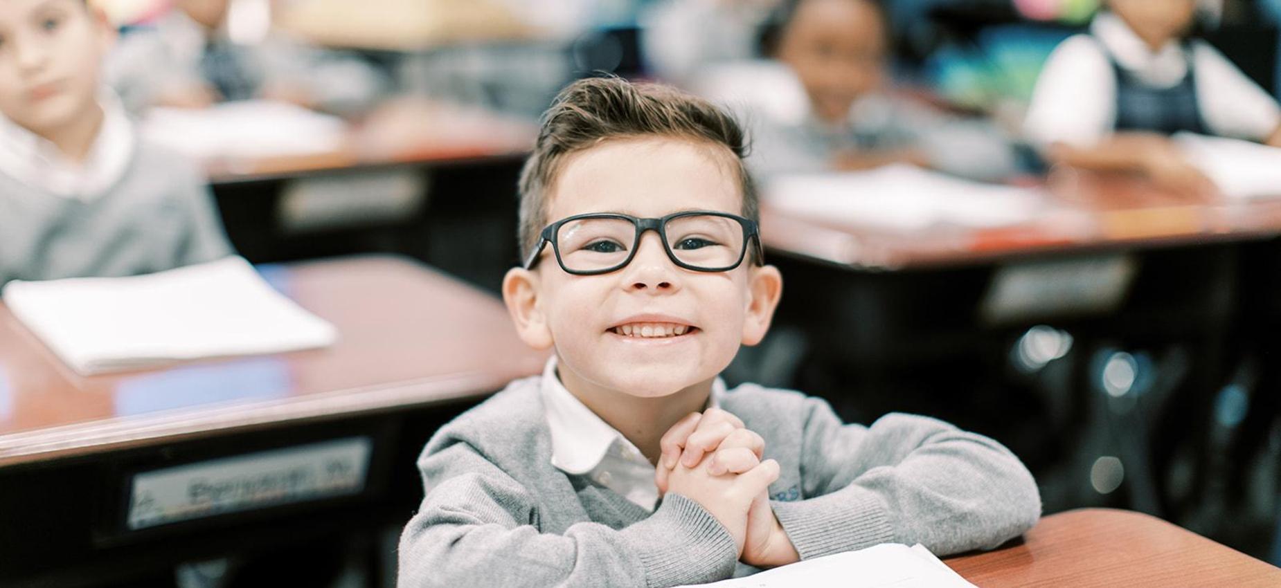 Smiling elementary student with glasses sits at his desk with his hands clasped