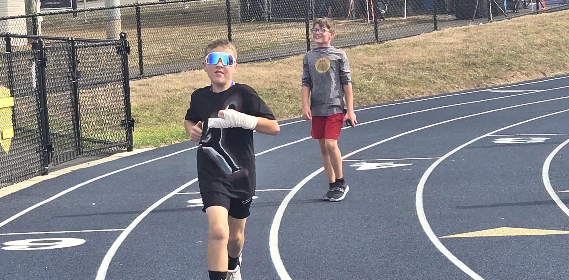 Boy running on a track with sunglasses and a bandaged hand, another boy behind him.