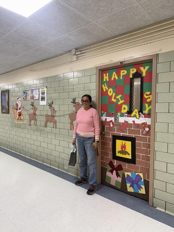 Ms. De La Cruz beside her decorated door. Santa on his sleigh with all his deer going are posted on the wall heading toward the door covered in gifts.
