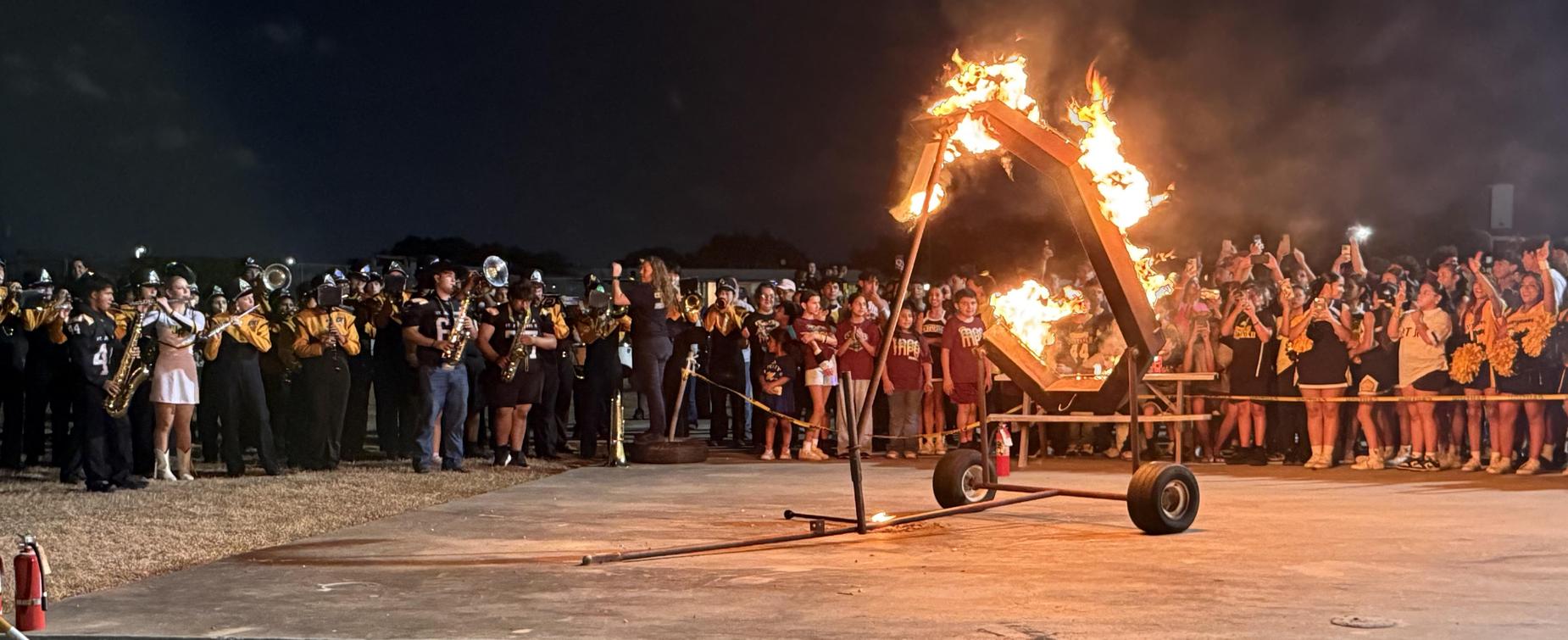 A lively crowd cheers as a large fire burns on a basketball court during an event.