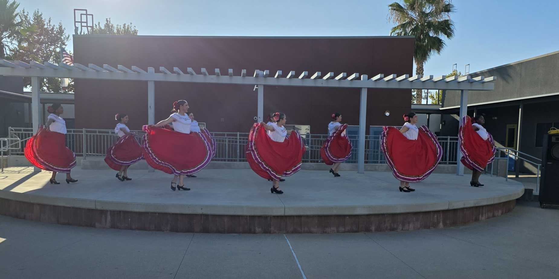 Dancers in red dresses performing on an outdoor stage with palm trees in the background.