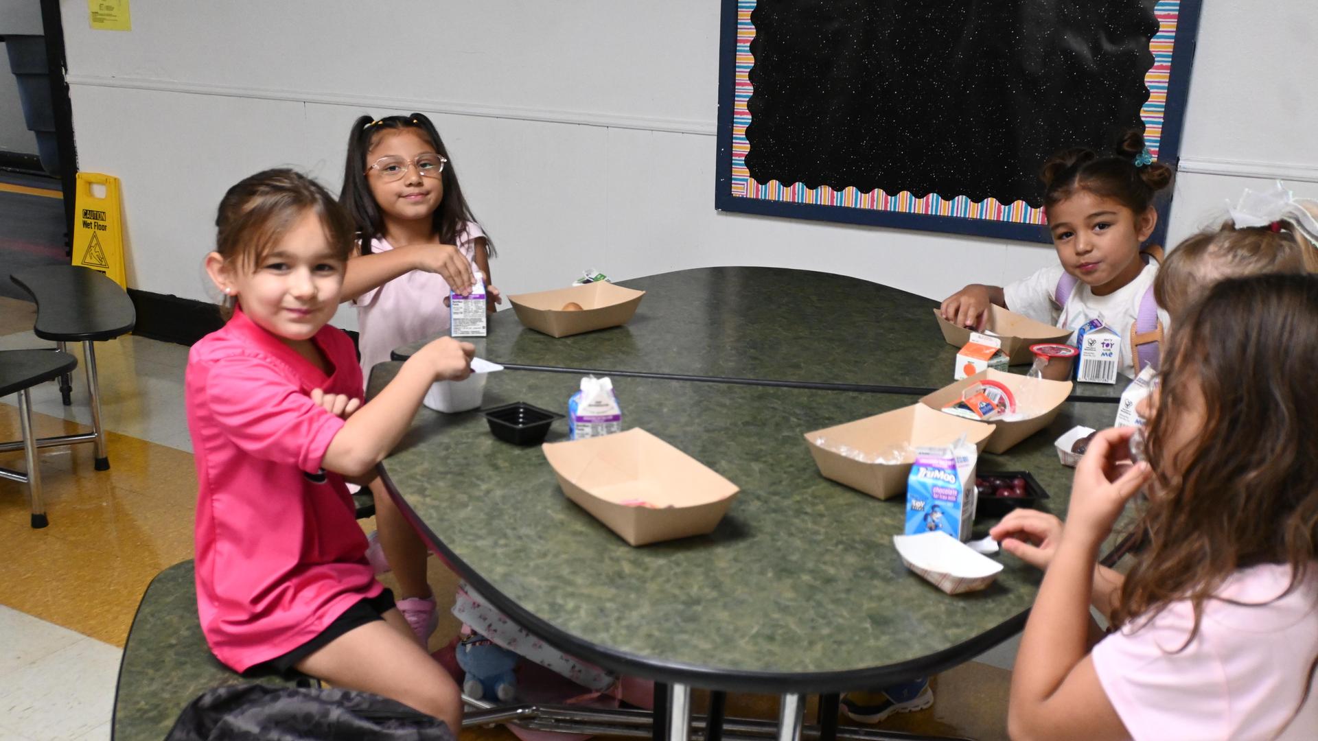 Children sitting at a table enjoying lunch with food trays and drinks.