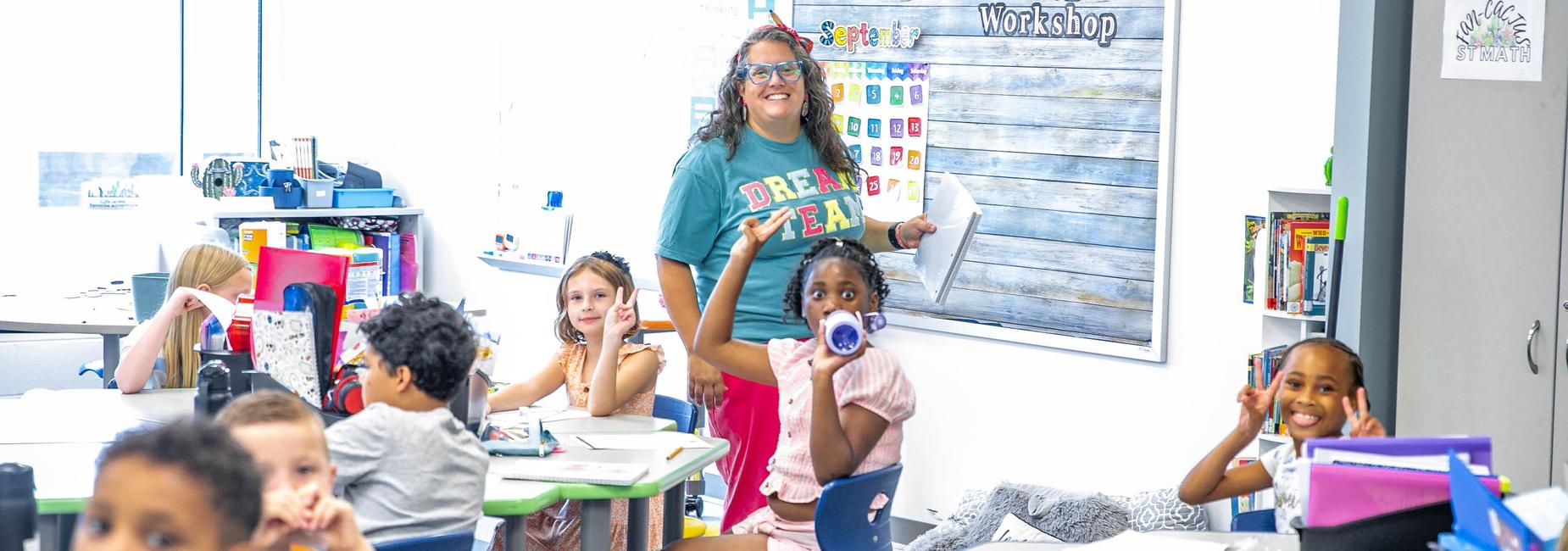 Teacher smiling and holding papers in a colorful classroom with students engaged.