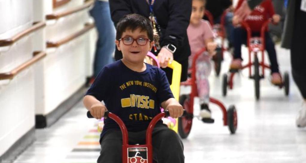 A young boy riding a tricycle in a bright school hallway filled with other children.