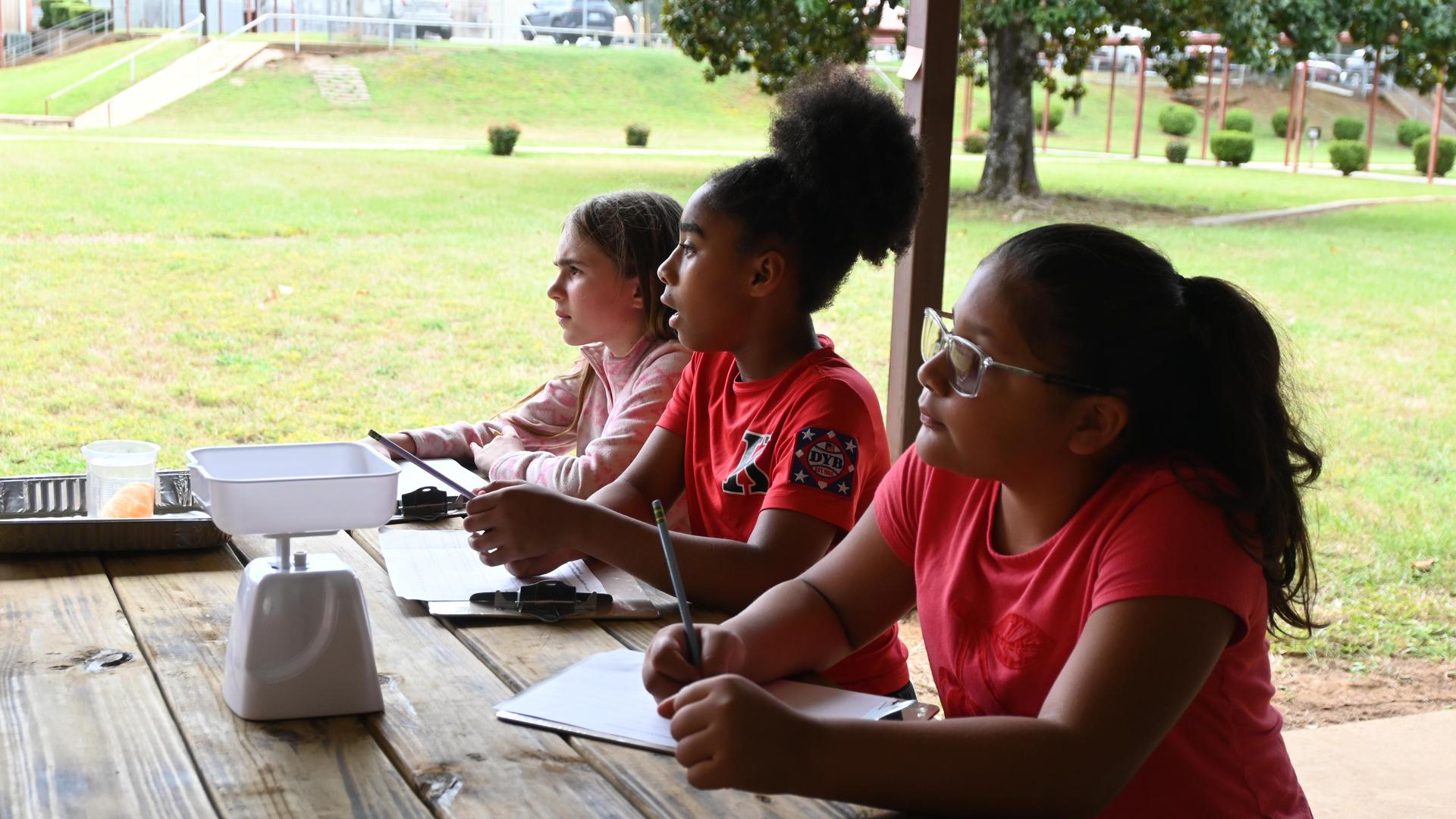 Three children sitting at a table, engaged in an outdoor learning activity.