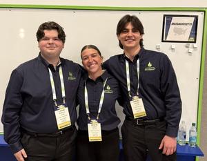 Three Taunton High School students wearing matching Massachusetts ProStart shirts and competition badges stand together smiling in front of a whiteboard.