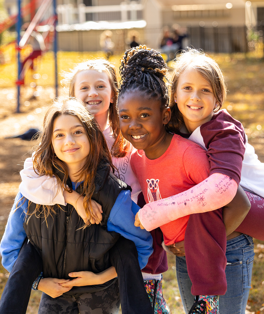 Four young girls smiling together outdoors on a playground, two of them giving piggyback rides to the others.