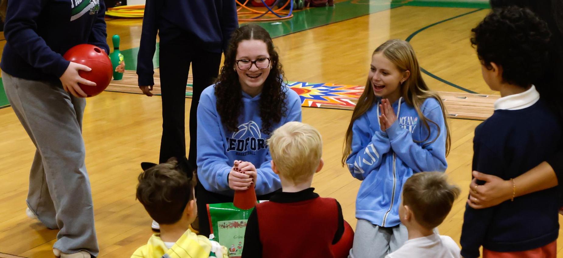 A group of children interacting with a smiling adult in a gymnasium setting.