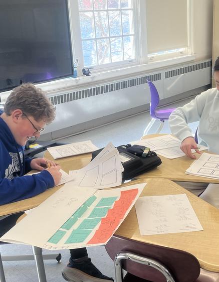 Two students working at a table with papers and a laptop in a classroom setting.