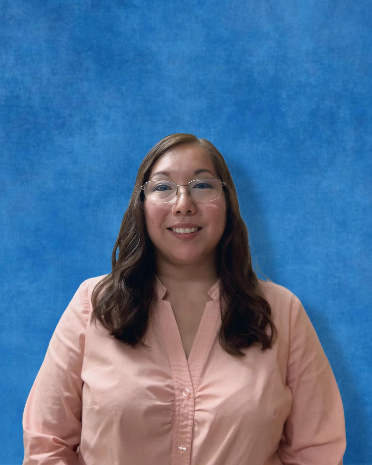 A woman with long brown hair, glasses, and a light pink blouse smiles in front of a blue background.