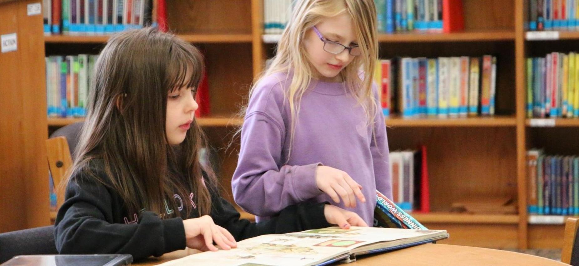 Two girls reading a book at a library table.