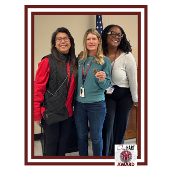 Three women smiling together, holding an award in front of an American flag.