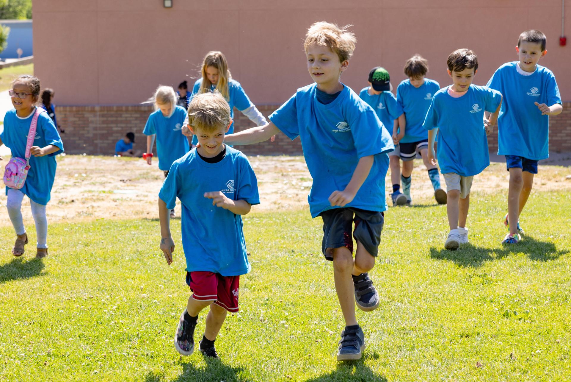 Children in matching blue shirts run across a sunny field toward the camera.