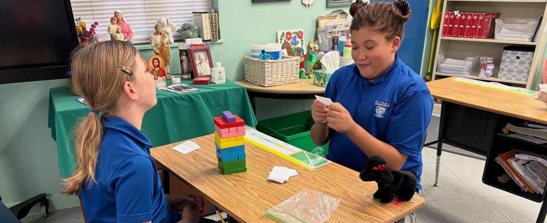Two students at a table playing a board game with colorful blocks and cards.