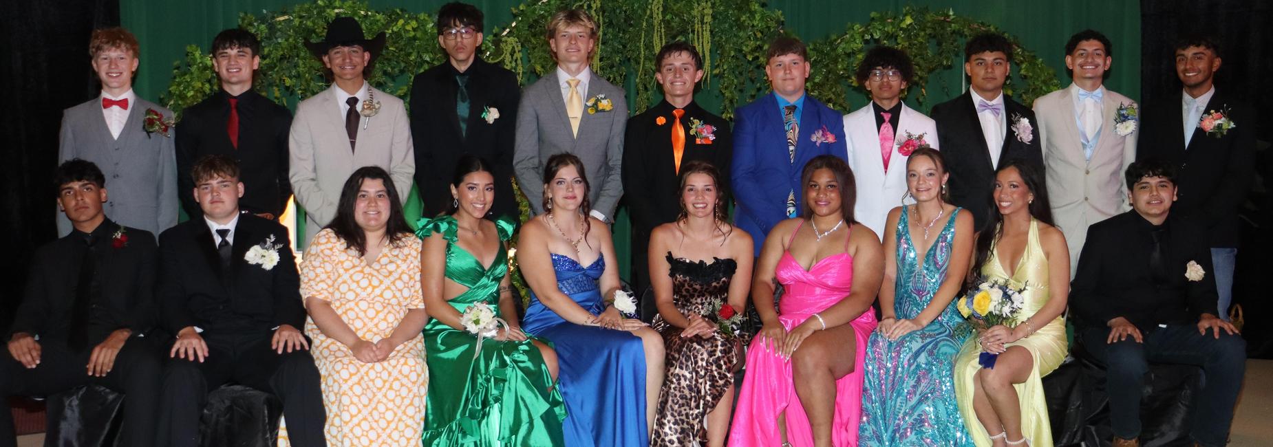Large group of teens in colorful formal wear, posing together with greenery backdrop.