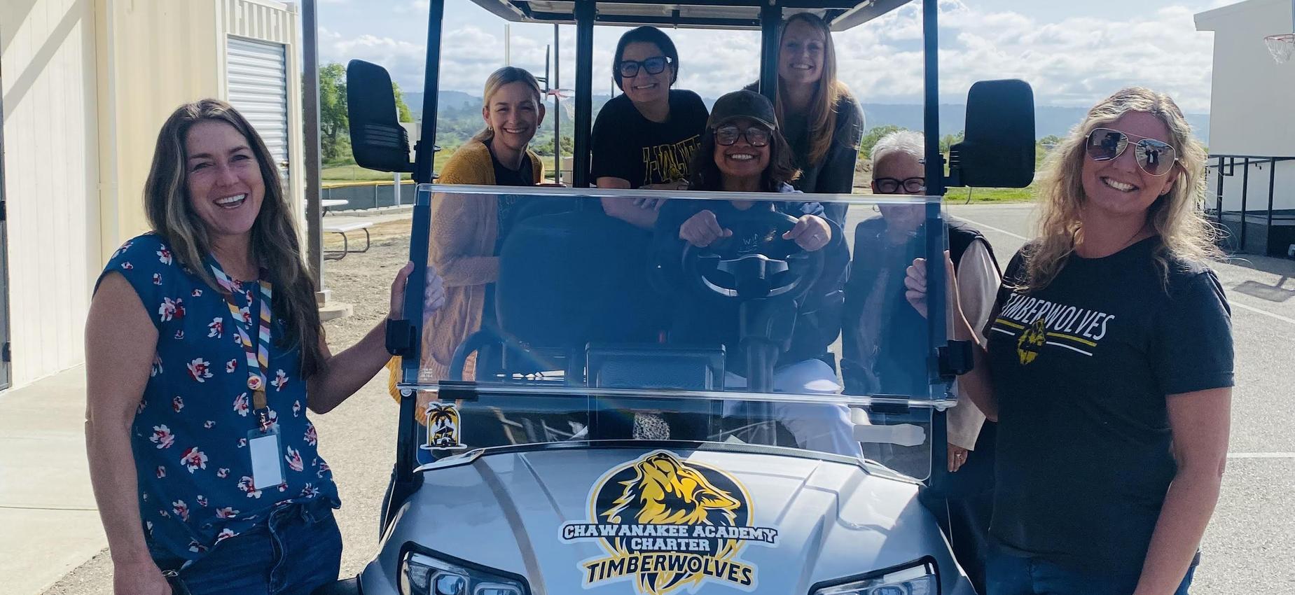 Group of smiling women and one man posing with a golf cart in daylight.