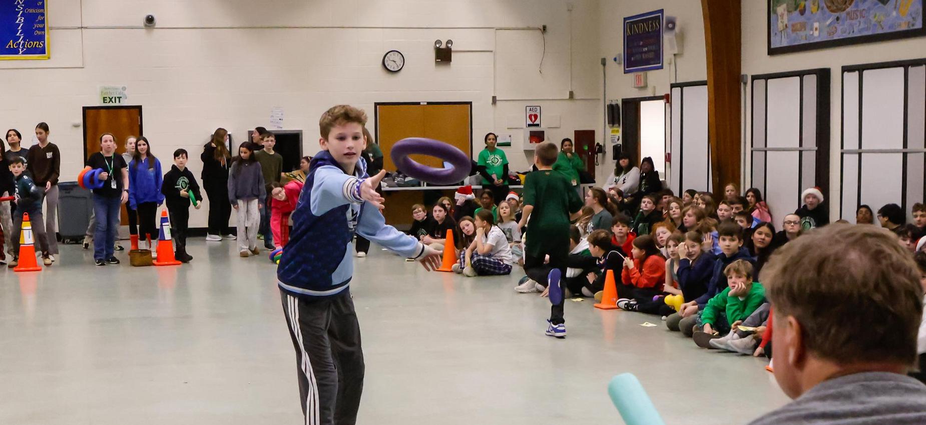 A student throws a ring towards another student during a school event with cheering classmates.
