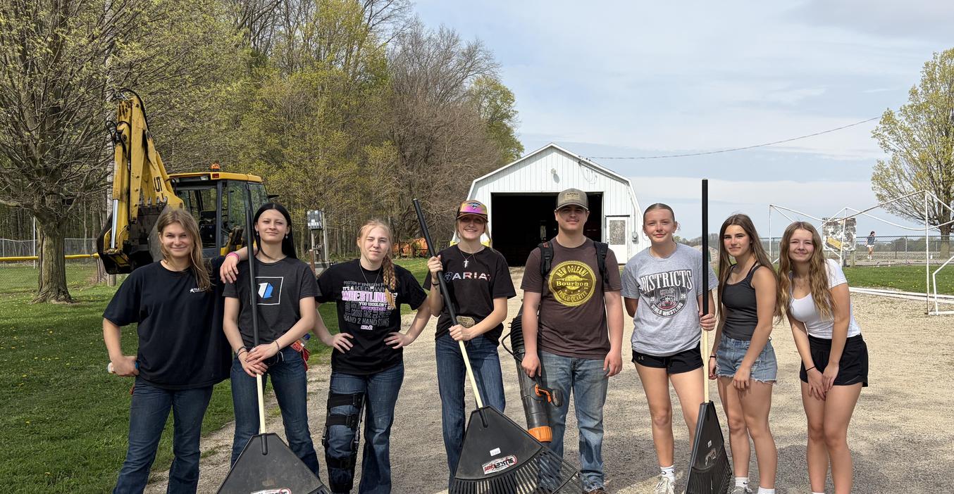 Group of eight young people holding brooms outdoors by a building.