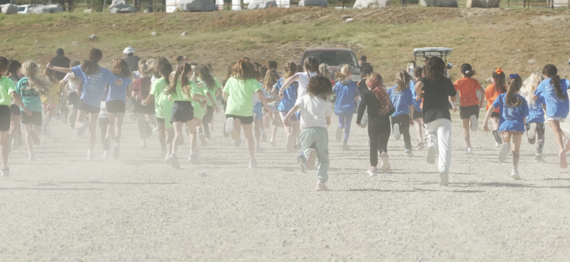Children running on a dusty path during a race, surrounded by grass and vehicles.