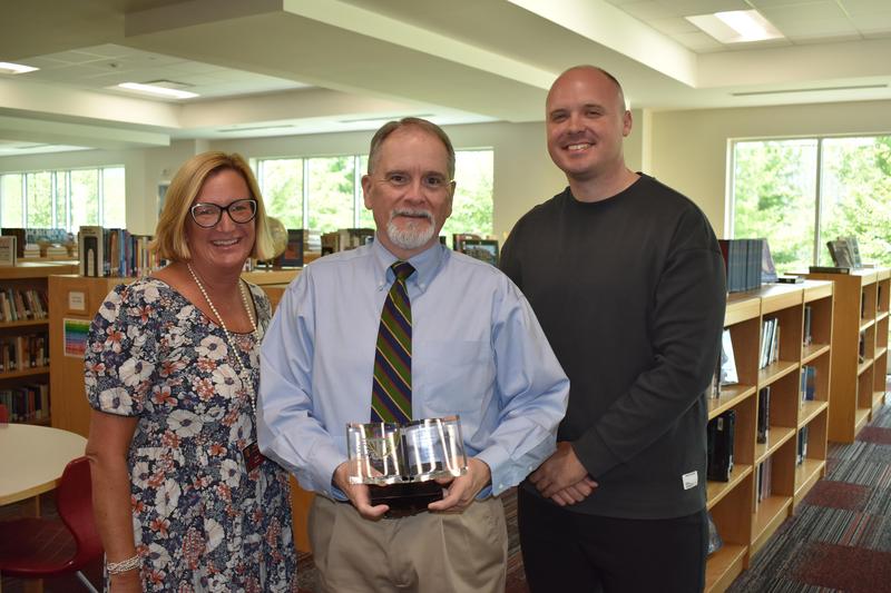 Three TJCA staff members pose in the Upper Campus library as the award recipient holds a crystal book-shaped plaque.