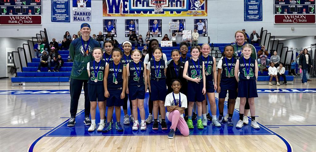A youth basketball team poses together on the court, smiling in their uniforms.