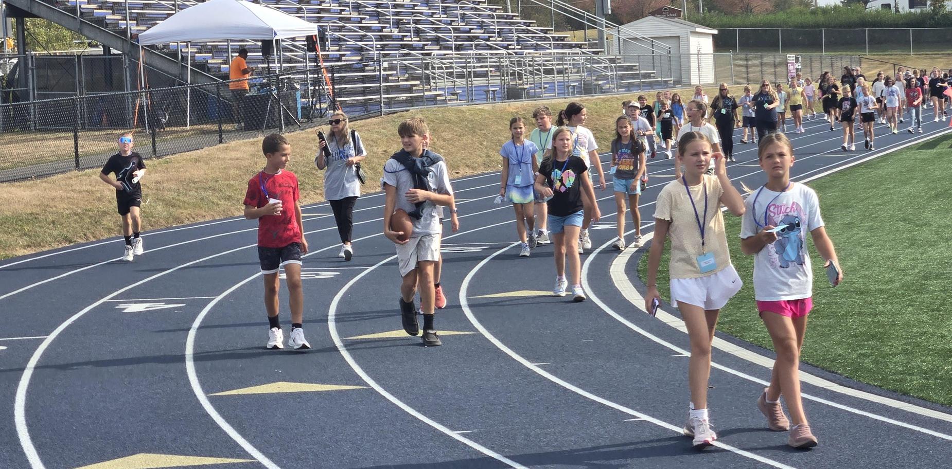 A crowd of children walking on a track, some holding items, with bleachers nearby.
