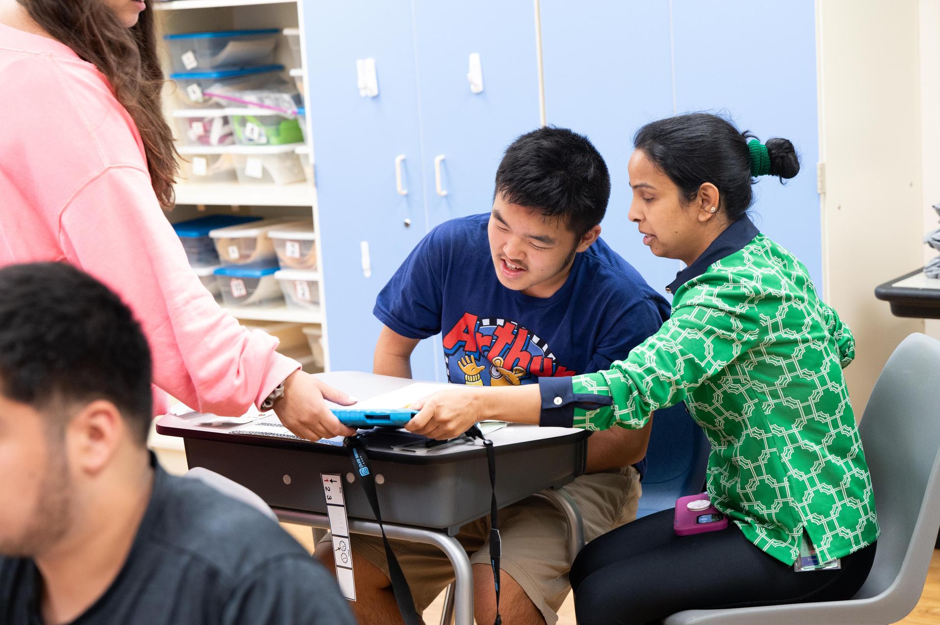 Student at desk wearing blue shirt teacher with green shirt helping.