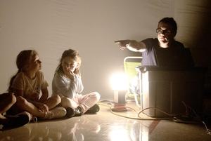 Adult pointing while sitting near a light source, with children listening intently.