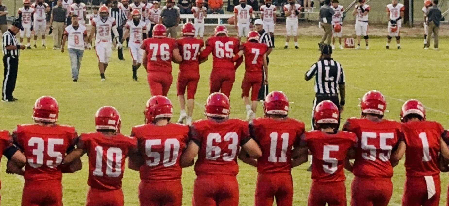 High school football players in red jerseys walking onto the field with spectators in background.