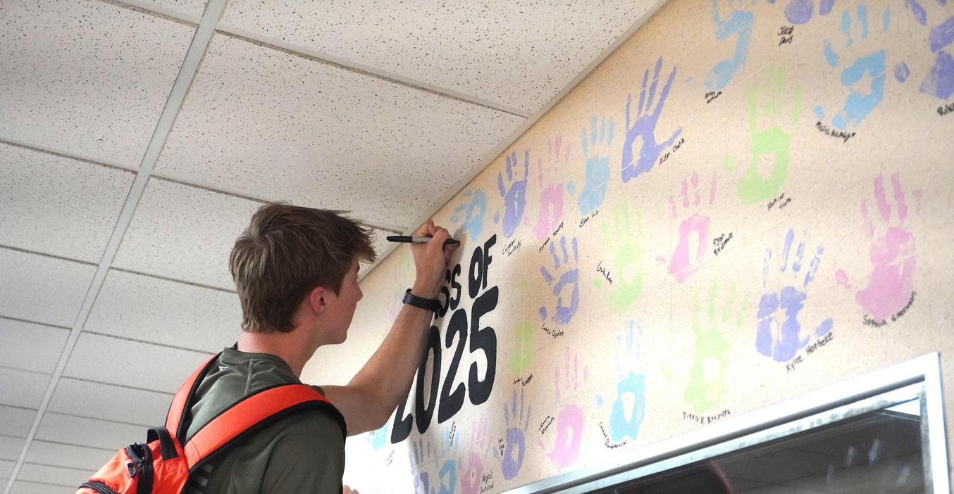 A senior writes his name under his handprint on the senior wall in the high school.