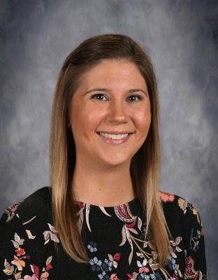 Smiling woman with shoulder-length hair in a black top with floral design against a gray backdrop.