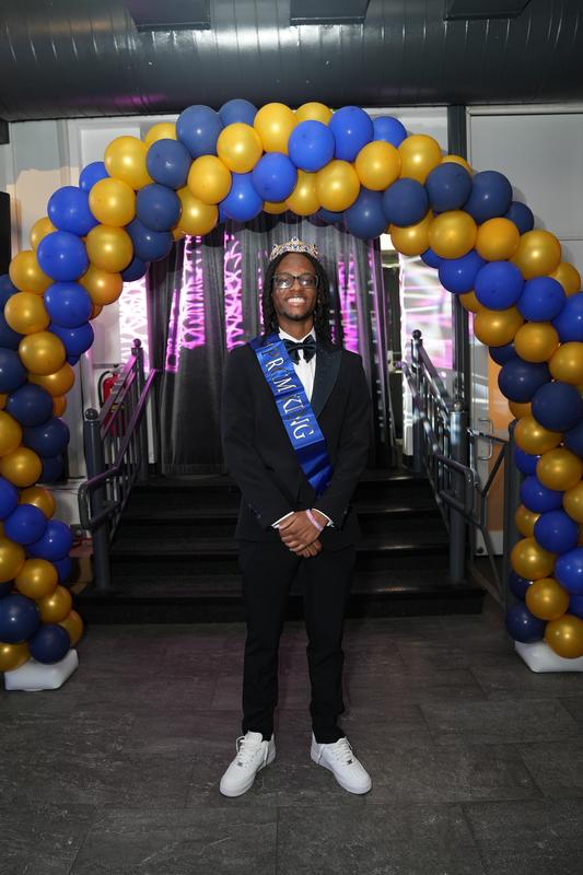 A person in a tuxedo stands under a balloon arch.