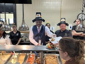 Principal McCaskill and foodservice staff serve food at Friendsgiving wearing pilgrim hats
