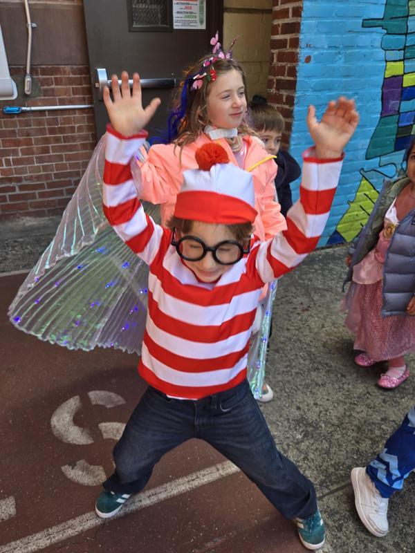 Boy dressed as Waldo with a striped shirt and had in red and white.  He is raising both his hands and wearing black large glasses.