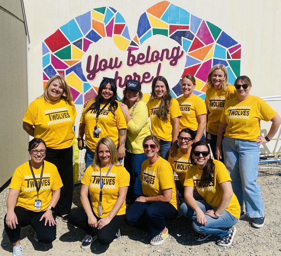 A group of twelve women in matching yellow shirts with 'TWOLVES' printed on them, smiling in front of a colorful mural.