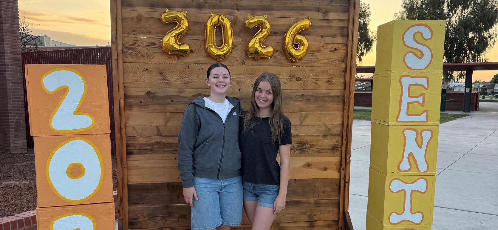 Two smiling girls pose in front of a wooden backdrop with balloons and painted blocks.