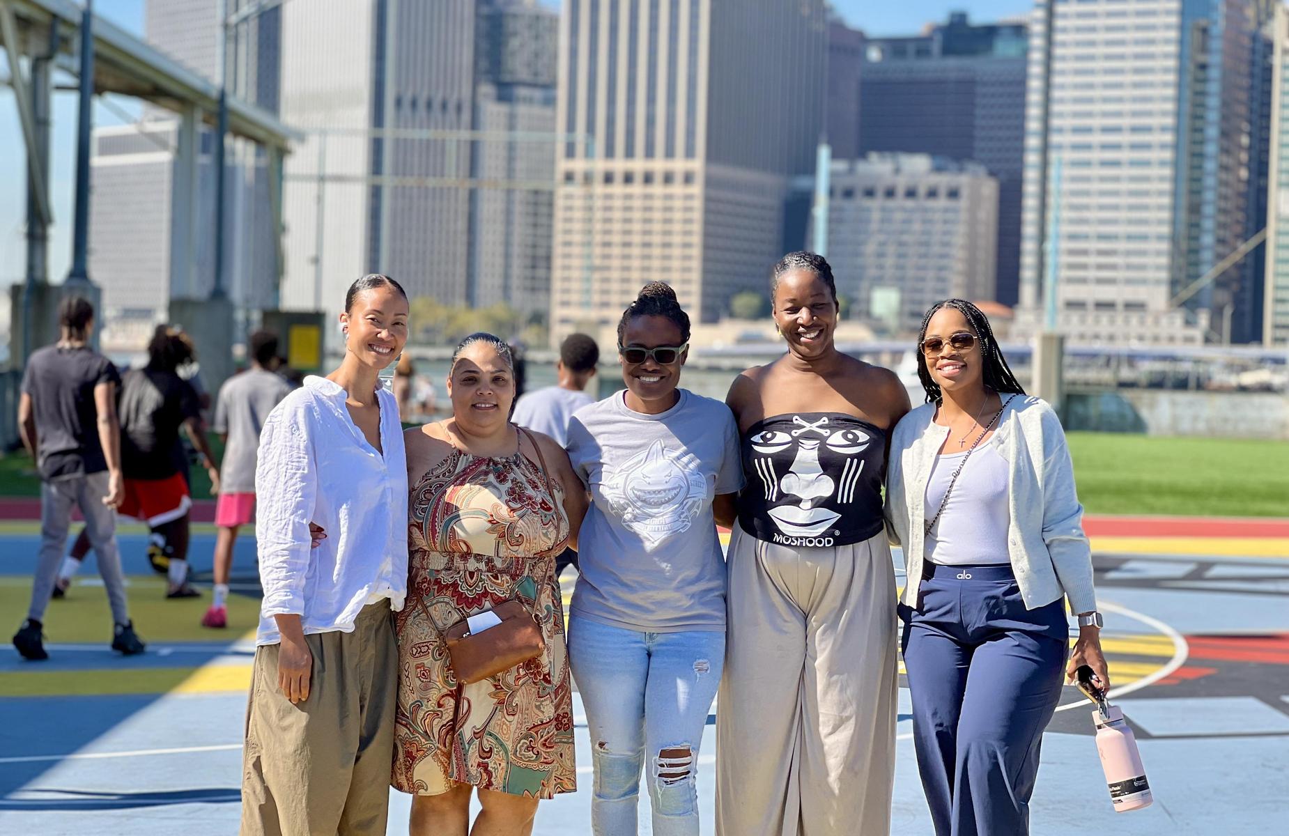 Five women posing together in an urban park with a city skyline in the background.