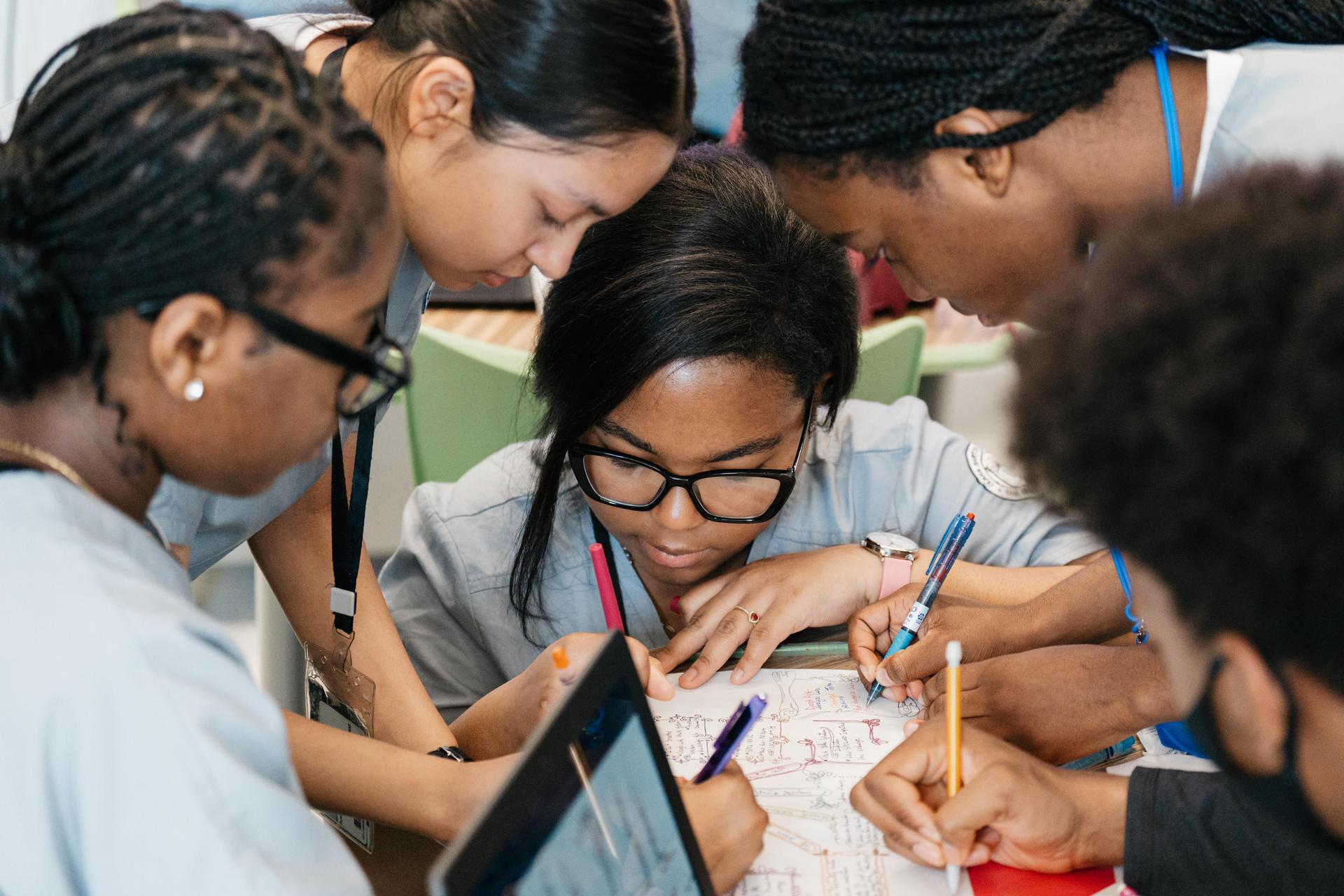 Students gather around a desk, collaborating and writing on a shared classroom assignment.