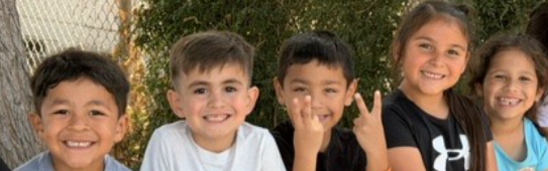 Five kids sitting together, smiling and making peace signs against a natural background.