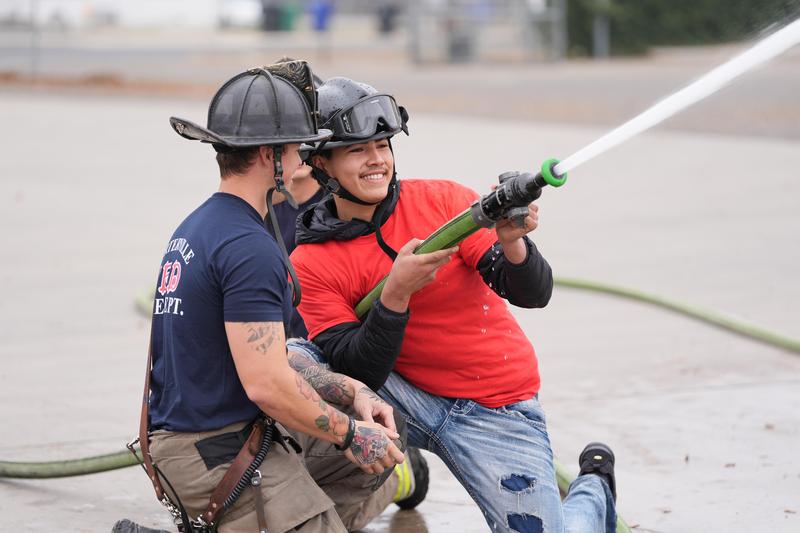 A smiling student in a red shirt and helmet operates a fire hose under the guidance of a firefighter wearing Porterville Fire Department gear. Water sprays from the hose as part of a hands-on fire training experience.