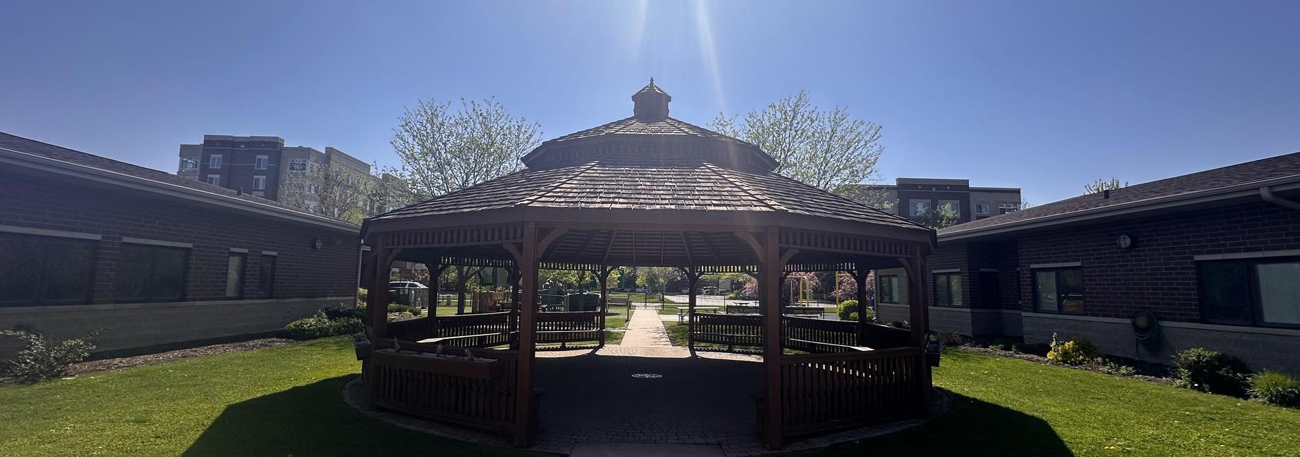 A wooden gazebo surrounded by green grass and trees under a clear blue sky.