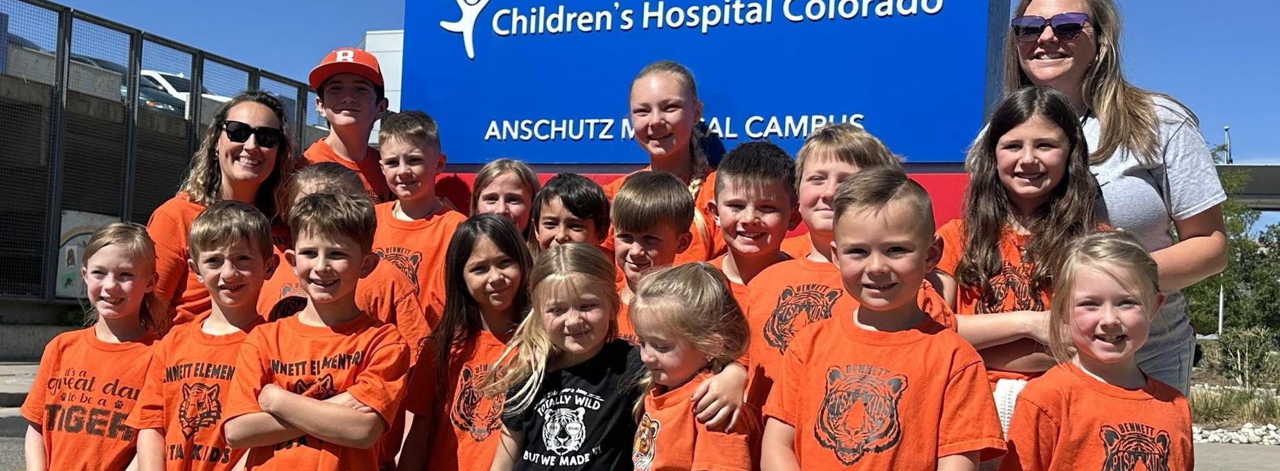Group of children and adults in orange shirts smiling in front of a sign at a medical campus.