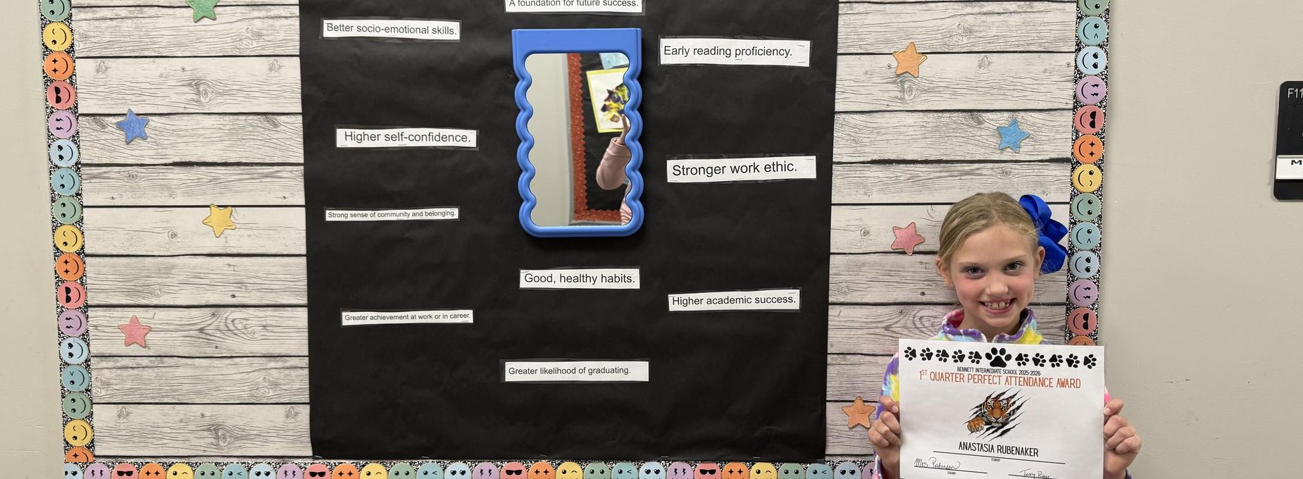 A girl stands with a certificate next to a bulletin board highlighting positive qualities.